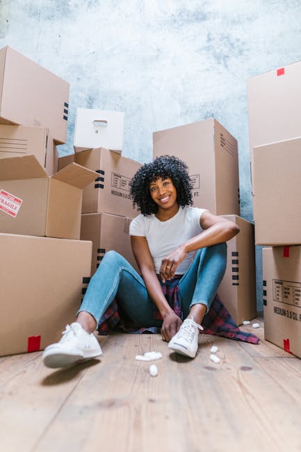 A young woman with curly black hair, wearing a white T-shirt, blue jeans, and white sneakers, is sitting on a wooden floor surrounded by multiple cardboard boxes of various sizes, some sealed with packing tape and others partially open. She is smiling and appears relaxed during a home relocation process. The boxes are stacked against a textured light blue wall, and some are marked with labels or fragile stickers, indicating they are ready for packing or transport. The scene is indoors, with natural or diffused lighting illuminating the space, and the woman is seated near the doorway or entrance area, suggesting a packing phase for a house move. This visual aligns with furniture transport and packing activities involved in house removals, as handled by companies like Man With a Van New Cross, supporting the moving process seamlessly.