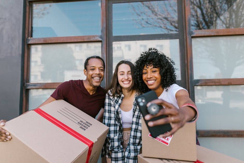 Three individuals standing outside a property, smiling and taking a selfie with a smartphone. They are holding cardboard boxes secured with red packing straps, indicating they are in the process of moving. The person on the left is holding a box labeled 'home moving supplies,' while the person on the right is also holding a box. The woman in the middle is smiling broadly, with her hand resting on a box. Behind them, large glass windows reflect trees and buildings, suggesting a residential setting. The scene is well-lit with natural daylight, and the individuals are dressed casually, demonstrating a home relocation scenario. This image relates to furniture transport, packing, and the logistics of house removals, often provided by companies like Man With a Van New Cross during estate moves, aligning with the topic of Amersham Estate moving tips for New Cross tenants.