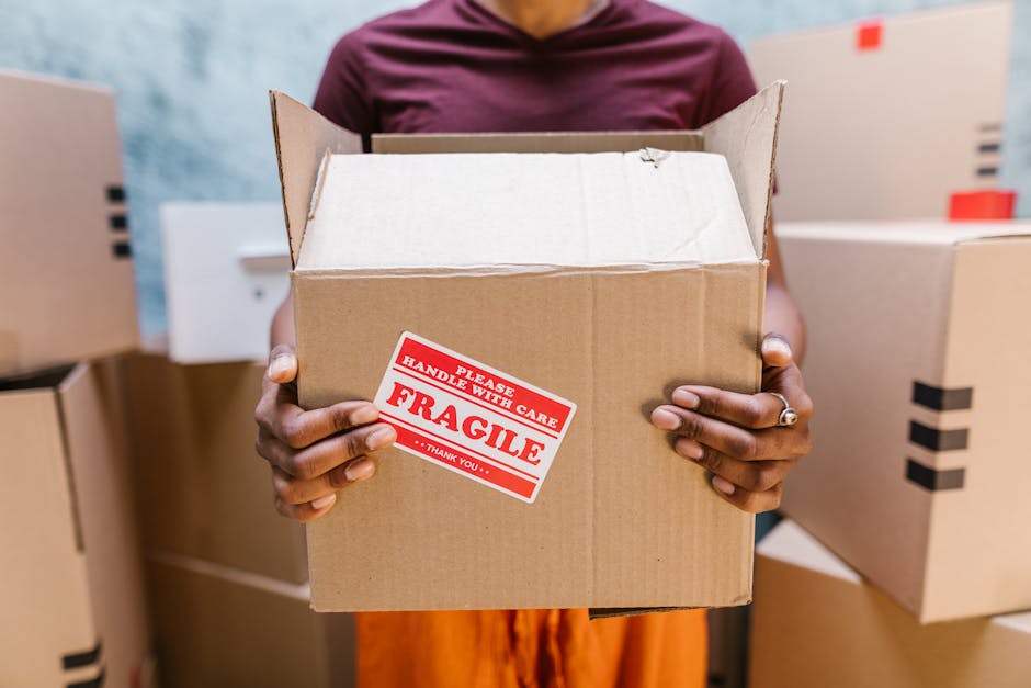A young woman with curly black hair, wearing a white T-shirt, blue jeans, and white sneakers, is sitting on a wooden floor surrounded by multiple cardboard boxes of various sizes, some sealed with packing tape and others partially open. She is smiling and appears relaxed during a home relocation process. The boxes are stacked against a textured light blue wall, and some are marked with labels or fragile stickers, indicating they are ready for packing or transport. The scene is indoors, with natural or diffused lighting illuminating the space, and the woman is seated near the doorway or entrance area, suggesting a packing phase for a house move. This visual aligns with furniture transport and packing activities involved in house removals, as handled by companies like Man With a Van New Cross, supporting the moving process seamlessly.