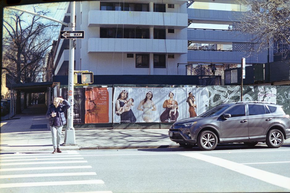 A street scene in New Cross showing a modern multi-storey building behind a construction site with a black temporary fence displaying large posters of four women in casual clothing. In the foreground, a woman wearing a dark jacket, light-colored pants, and a beanie is standing on a pedestrian crossing, holding a phone and adjusting her hood. A dark grey SUV is parked on the street next to the curb, partially blocking the view of the sidewalk. The area includes a street sign indicating one way, a traffic light mounted on a pole, and trees with bare branches suggesting winter or early spring. The scene captures the urban environment typical for house removals or furniture transport, with visible elements like parking, pedestrian access, and building surroundings that might be involved in a home relocation or moving process as managed by companies like Man With a Van New Cross.