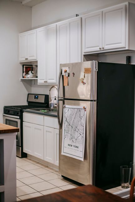 The image depicts a kitchen interior with white upper and lower cabinets, a stainless steel refrigerator with various notes and a map attached to its front, and a black oven and stove situated beneath the cabinets. The countertops are light-colored, and a sink with a faucet is visible next to the refrigerator. A small microwave is placed inside an open cabinet above the stove, and the kitchen floor is tiled with light-colored square tiles. There is a wooden table partially visible in the foreground, and the lighting appears to be natural, illuminating the space evenly. This setting is indicative of a home environment preparing for packing and moving activities, with furniture and appliances ready for transport by a professional removals service such as Man With a Van New Cross.