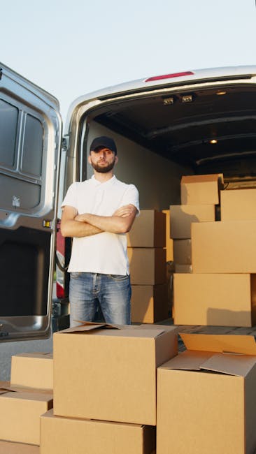 A man with a beard, wearing a black cap, white t-shirt, and jeans, stands with arms crossed in front of the open rear doors of a van parked outdoors on a paved surface. Inside the van, several large, 