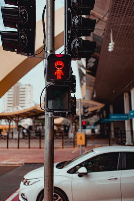 A pedestrian traffic light showing a red figure indicating 'stop' is mounted on a metal pole, which also supports multiple black traffic signals with wires attached. In the background, an urban street scene features a white car parked close to the curb, with buildings and a covered outdoor seating area visible under a modern canopy structure. The scene is taken during daylight hours, with a partly cloudy sky visible in the distance. The image captures a typical city environment relevant to home relocation logistics, with clear visuals of traffic controls and vehicle positioning, illustrating the structured flow of urban transport and movement that can be involved during furniture transport and moving services, as provided by companies like Man With a Van New Cross.