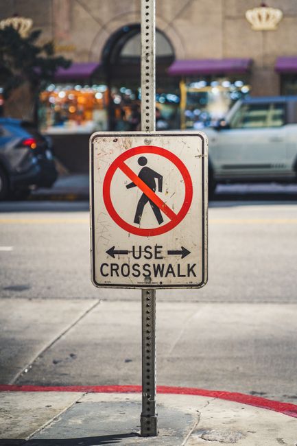 A rectangular metal sign mounted on a vertical pole on a sidewalk in an urban area, displaying a red circle with a diagonal line crossing out a black silhouette of a person walking, indicating no pedestrian access. Below, the sign reads 'USE CROSSWALK' with black arrows pointing left and right, directing pedestrians to cross at designated crossings. In the background, blurred vehicles are parked along the street, with storefronts featuring purple awnings and illuminated signage. The surrounding environment is well-lit, suggesting evening or night time, with a mix of natural and artificial lighting. This sign is positioned near the entrance of a property, which may be part of a home relocation or moving process, as handled by Man With a Van New Cross, a professional removals company specializing in house moves and furniture transport in the SE14 area. The setting emphasizes urban safety and logistics during a home or furniture moving activity.