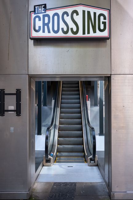 A set of escalators inside a building with a metal frame and black rubber steps, seen through an open doorway beneath a sign that reads 'The Crossing.' The space surrounding the escalators includes metallic walls and a small black mat on the floor at the entrance, indicating a station or transit point. The image highlights the interior environment where home relocation or moving services provided by Man With a Van New Cross may involve navigating stairs or access points during furniture transport or packing and moving processes, illustrating the logistical considerations of house removals within a busy urban setting.