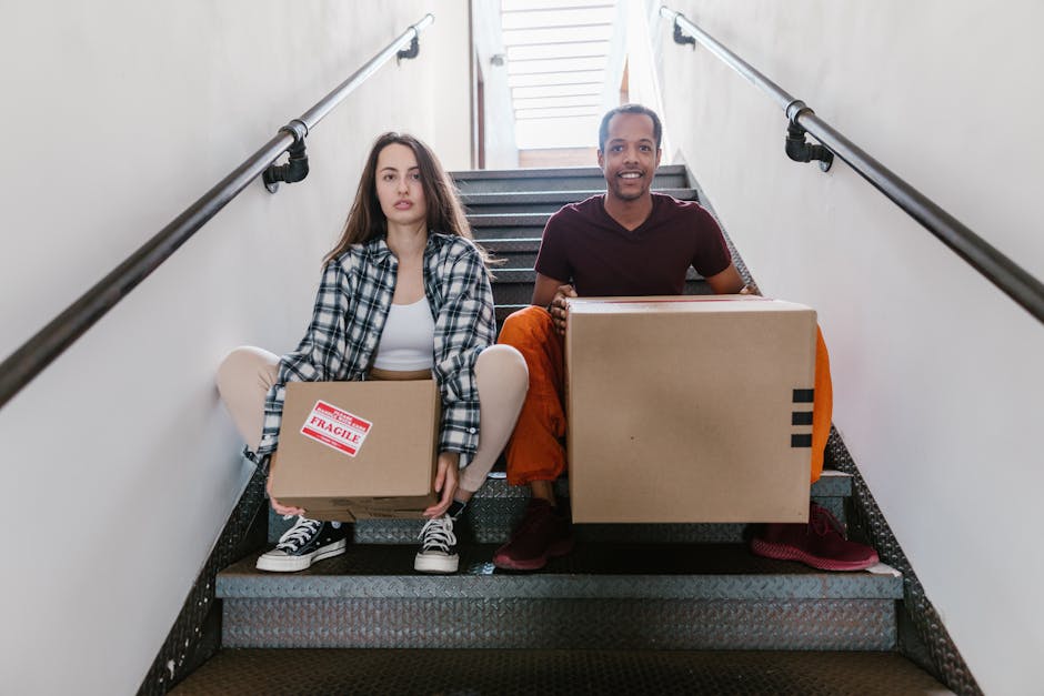 Two movers from Man With a Van New Cross are seated on a staircase inside a property, preparing to transport boxes during a home relocation. The woman on the left holds a small cardboard box marked 'Fragile' with packaging tape and is dressed in a checkered shirt, white top, and sneakers. The man on the right, wearing a maroon t-shirt and orange trousers, carries a larger cardboard box with black tape. Both are positioned at the bottom of a staircase with metal railings on either side, leading upwards to a well-lit area with a window at the top. The surroundings include a plain white wall, and the staircase is carpeted with a dark, textured material. The image captures the loading process involved in furniture transport and packing during house removals, illustrating the careful handling and packing of household items typical in professional moving services like those offered by Man With a Van New Cross.