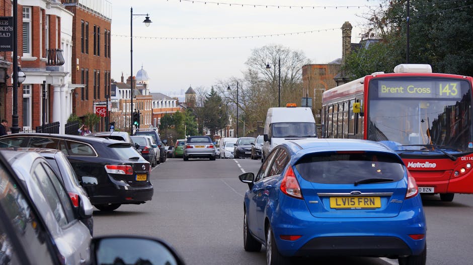 A busy street scene in New Cross with multiple parked cars, including a black hatchback, a silver estate, and a blue hatchback, lining both sides of the road. A red Metroline bus displaying route number 143 to Brent Cross is passing through and partially occupying the right side of the image. In the background, white and brick residential buildings with traditional architecture are visible, along with trees and lamp posts along the sidewalk. The scene is captured during daylight with overcast weather, presenting a typical urban environment suitable for house removals and furniture transport logistics. The image illustrates the typical street context where Man With a Van New Cross may carry out home relocation or packing and moving activities, with parked vehicles and moving elements seamlessly integrated into the scene.