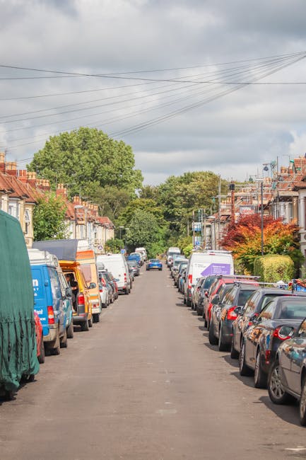 A wide residential street with parked cars lining both sides of the road, including hatchbacks, saloons, and vans, some covered with protective tarps and plastic wraps. The street features neatly maintained terraced houses with small front gardens, some with trees and flowering shrubs, and overhead power lines stretch across the cloudy sky. The pavement is visible on either side, and in the distance, larger trees and greenery can be seen beyond the rooftops. The scene suggests a typical urban neighbourhood suitable for home relocation services, with [COMPANY_NAME] likely involved in the loading or unloading process of household furniture and packing materials, such as cardboard boxes and removals blankets, that are ready for transport. The natural daylight illuminates the scene, highlighting the organized parking and typical street layout used during local house moves in Lewisham or New Cross.