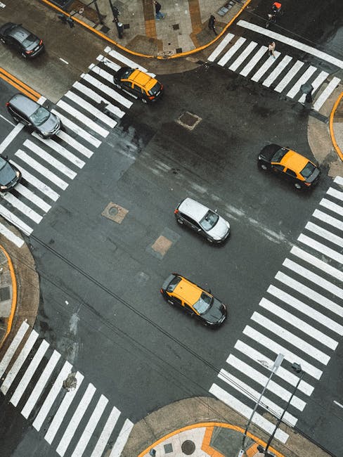 An aerial view of a city intersection showing multiple vehicles, including yellow and black taxis and a silver car, moving along wet asphalt with clearly marked pedestrian crossings and crosswalks. The surrounding sidewalk features a paved pedestrian area with orange tactile paving at the corners, and a few pedestrians are walking nearby. The scene captures the urban environment during overcast weather, with the street appearing shiny from recent rain. This image reflects the logistical aspects of home relocation and furniture transport, as part of the moving process, with visible road markings and vehicle traffic supporting efficient transportation for removals services offered by Man With a Van New Cross.