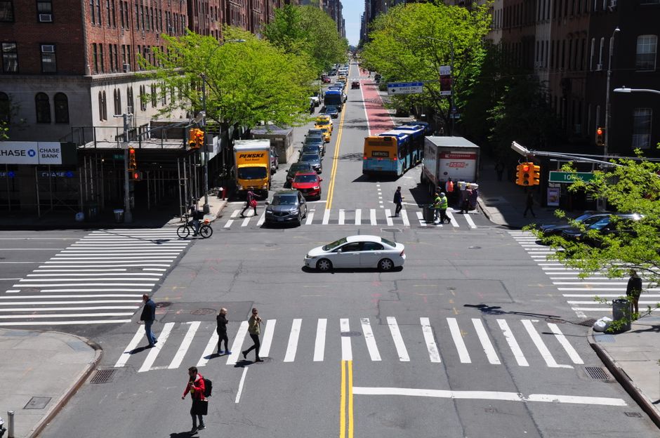 An aerial view of a city intersection showing multiple vehicles, including yellow and black taxis and a silver car, moving along wet asphalt with clearly marked pedestrian crossings and crosswalks. The surrounding sidewalk features a paved pedestrian area with orange tactile paving at the corners, and a few pedestrians are walking nearby. The scene captures the urban environment during overcast weather, with the street appearing shiny from recent rain. This image reflects the logistical aspects of home relocation and furniture transport, as part of the moving process, with visible road markings and vehicle traffic supporting efficient transportation for removals services offered by Man With a Van New Cross.