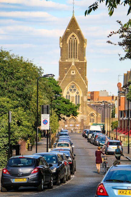 A narrow street in New Cross with parked cars lining both sides, including hatchbacks and sedans in various colours. The street is paved and features a pedestrian crossing with two individuals, one riding a bicycle and another walking, both dressed in casual clothing. Green trees with full foliage extend over parts of the street, providing shade, and lampposts are positioned along the pavement. In the background, a historic stone church with large arched windows and a tall, pointed steeple dominates the scene, set against a partly cloudy sky. This setting illustrates typical urban surroundings where house removals and furniture transport may encounter narrow streets and parking considerations, such as those discussed in the New Cross Gate moving checklist by Man With a Van New Cross.