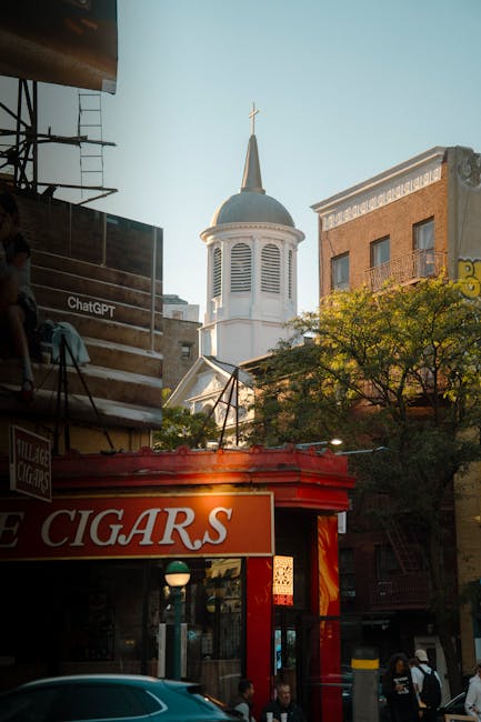 A narrow residential street in New Cross with tightly parked cars lining both sides of the asphalt roadway, including various models and colours; some vehicles are covered with protective fabric or plastic sheeting. The street features terraced houses with brick facades, small front gardens, and front doors facing the pavement. Overhead, there are power lines and a cloudy sky, suggesting a daytime setting. In the context of home relocation or furniture transport, this street layout presents typical parking and access challenges for moving services such as those offered by Man With a Van New Cross, highlighting the importance of careful planning during a house move on narrow streets with limited parking options.