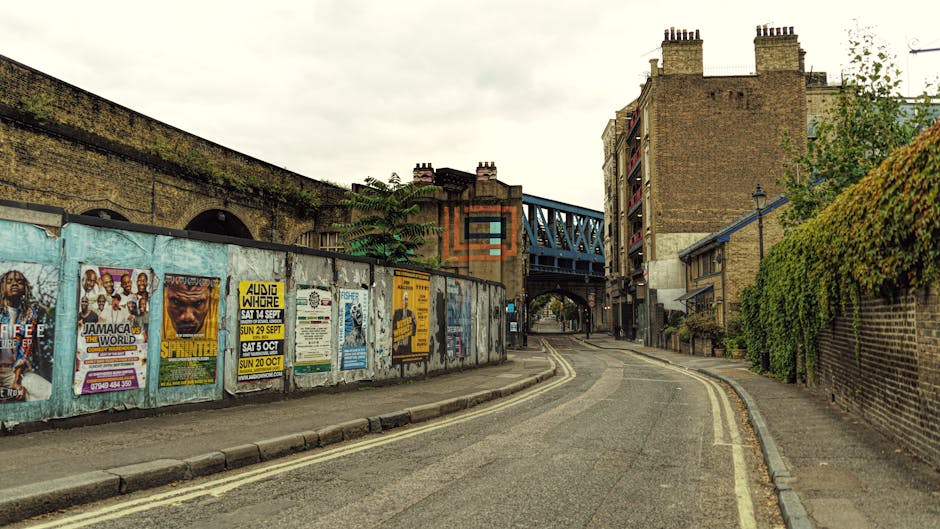 A narrow cobblestone street in a residential area with historic brick and stone buildings on either side, some with large windows and decorative facades. The street is lined with double yellow lines o