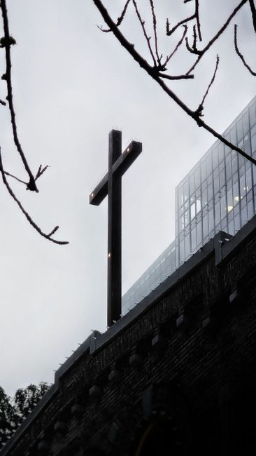 A black metal cross mounted on the edge of a building rooftop, with a modern glass structure visible behind it, under an overcast sky. In the foreground, bare tree branches extend into the frame, suggesting a winter or late autumn setting. The image captures the exterior of a religious or memorial site, with the cross as a prominent feature. The scene appears to be on the roof of a building involved in home or commercial property relocation, with part of the roof's brickwork and the glass structure indicating urban surroundings. This setting aligns with the context of home relocation or moving services provided by Man With a Van New Cross, showcasing a moment of loading or setting up a property in a city environment.