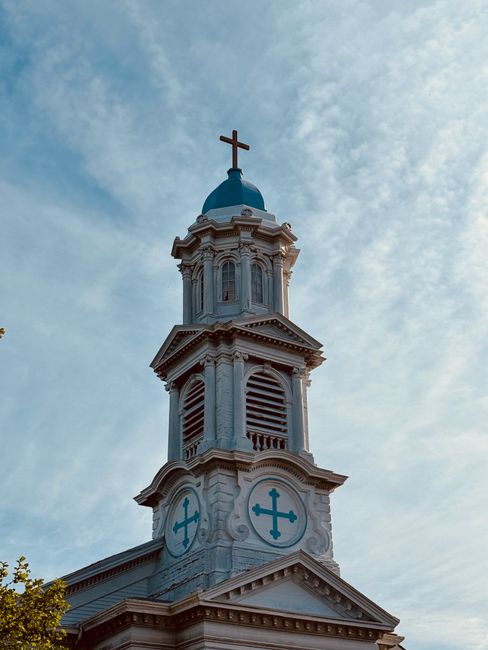 A black metal cross mounted on the edge of a building rooftop, with a modern glass structure visible behind it, under an overcast sky. In the foreground, bare tree branches extend into the frame, suggesting a winter or late autumn setting. The image captures the exterior of a religious or memorial site, with the cross as a prominent feature. The scene appears to be on the roof of a building involved in home or commercial property relocation, with part of the roof's brickwork and the glass structure indicating urban surroundings. This setting aligns with the context of home relocation or moving services provided by Man With a Van New Cross, showcasing a moment of loading or setting up a property in a city environment.
