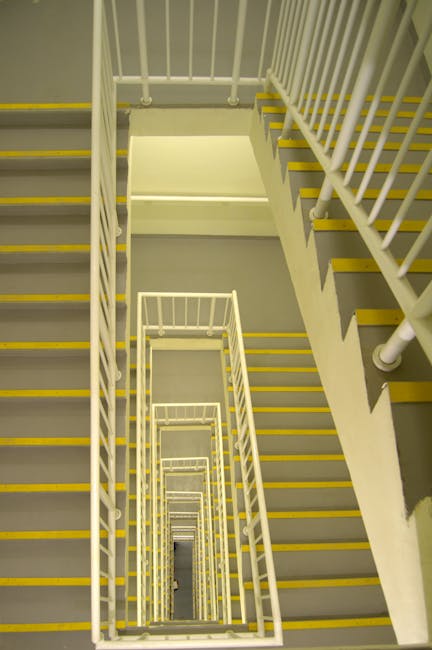 A view looking down a multi-storey staircase inside a residential building, showing a series of white metal railings with vertical bars and yellow safety strips along the edges of each step. The stairs are made of concrete with grey treads, inside a well-lit stairwell with neutral-colored walls. The image captures the descent through several levels, emphasizing the repetitive pattern of the staircase. This type of scene illustrates typical internal stairwell environments in flats, relevant to house removals and furniture transport during home relocation services provided by companies like Man With a Van New Cross, as depicted on their website page about moving down tight stairwells on SE14 flats.