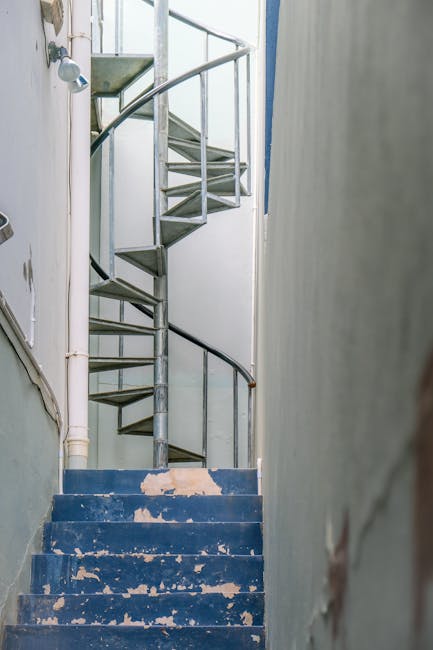 Interior view of a narrow staircase inside a residential building, with worn blue-painted steps showing chipped paint and signs of use. The staircase has a spiral metal structure with a black handrail on the outer side, leading upwards towards a small landing. To the left, a white wall features a small wall-mounted light fixture. Adjacent to the staircase, there is a white metal pipe running vertically alongside the wall. The surrounding environment appears aged but functional, with visible wear consistent with frequent use. The setting suggests a typical internal staircase in an older UK property, relevant for home relocation and furniture transport. In the foreground, a partially visible door frame indicates an entry point to the staircase area. This image is associated with the type of moving logistics, such as house removals performed by Man With a Van New Cross, specializing in moving down tight stairwells during house or flat relocations in SE14 and surrounding areas.