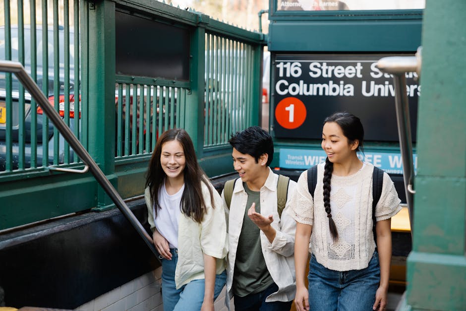 A group of three young people, two women and one man, are walking up a flight of outdoor stairs leading from an underground train station near Columbia University, in an urban area. The women have long dark hair, with one wearing a white cardigan over a white shirt and the other with a beige sweater, both carrying backpacks. The man, with short dark hair, is dressed in a light-colored jacket over a grey shirt and also carrying a backpack. They are smiling and appear engaged in conversation, with daylight illuminating the scene. Behind them is a green metal fence and the station signage indicating '116 Street Columbia University' and the bus number 1, suggesting they are preparing for a home relocation or move-in process. The environment includes visible pavement, a portion of a yellow taxi, and the entrance to the station, aligning with moving logistic activities supported by [COMPANY_NAME], such as packing and transport during a house or student move.