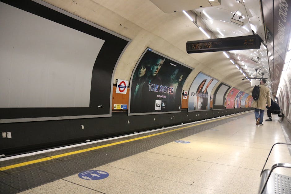 Inside a London Underground station, the curved platform wall is decorated with large advertisement posters, including one for the TV series 'The Sister.' A man in casual clothing with a backpack is walking along the platform near the edge, close to a yellow safety line, while another person is seated on a bench further down. The ceiling is illuminated with bright linear lighting fixtures, and digital signs hang overhead displaying train information such as 'Stokebridge Park' and 'Xlen 4 mins,' indicating scheduled train arrivals. The station features a modern concrete design with tiled flooring, and the atmosphere suggests it is part of a house or apartment relocation process, potentially involving a move from student accommodation near Goldsmiths to New Cross Station, supported by the professional services of Man With a Van New Cross. The scene captures typical moving logistics activity, including passenger transit and preparation for transportation, aligned with moving and furniture transport services.