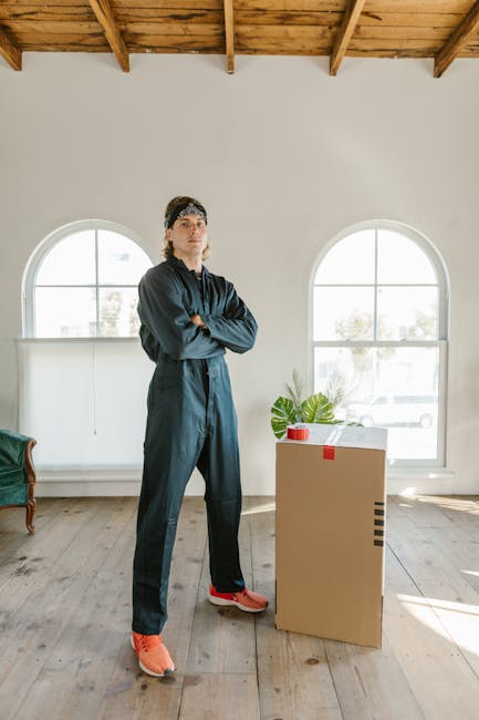 A man dressed in dark work clothing and orange shoes stands inside a spacious room with light-colored wooden flooring, wooden ceiling beams, and two large arched windows allowing natural light to fill the space. He is positioned next to a cardboard moving box that is sealed with red tape and contains a potted plant, which is placed on a small table or surface. The room appears to be prepared for a home relocation or packing process, with minimal furniture visible, and the natural lighting highlights the clean, organized environment. This setting exemplifies the initial stages of furniture transport and packing in a house move, consistent with services provided by Man With a Van New Cross specialized in removals and moving logistics.