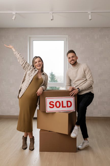 A couple stands inside a modern, minimally decorated room with light-colored wallpaper and a large window, celebrating the sale of their home. The woman, wearing a beige checkered shirt and a gold dress, points happily towards the window with her right arm raised, while the man, dressed in a cream sweater and dark pants, holds a cardboard box labeled 'SOLD' and leans on a stack of three additional cardboard boxes. The boxes are arranged on the wooden floor, indicating a recent or upcoming move as part of a house relocation process. Natural light floods the room from the window behind them, enhancing the bright, neutral environment. This image relates to property sales and the logistics of home removals, demonstrating the completion of a property transaction and the subsequent packing and transport activities handled by services like Man With a Van New Cross.