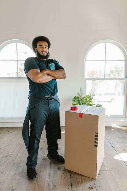 A man dressed in dark work clothing and orange shoes stands inside a spacious room with light-colored wooden flooring, wooden ceiling beams, and two large arched windows allowing natural light to fill the space. He is positioned next to a cardboard moving box that is sealed with red tape and contains a potted plant, which is placed on a small table or surface. The room appears to be prepared for a home relocation or packing process, with minimal furniture visible, and the natural lighting highlights the clean, organized environment. This setting exemplifies the initial stages of furniture transport and packing in a house move, consistent with services provided by Man With a Van New Cross specialized in removals and moving logistics.